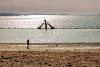 Along the seafront at Plage de Bon-Secours in Saint-Malo, Brittany, a solitary woman walks, the sea whispering its quiet memories as the breeze carries them away (zilverbat.)